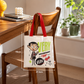 A white canvas tote bag with red handles hangs from a wooden chair in a bright room. The bag has a cartoon character print and the words 'VERY IMPORTANT CHAP BEAN'.