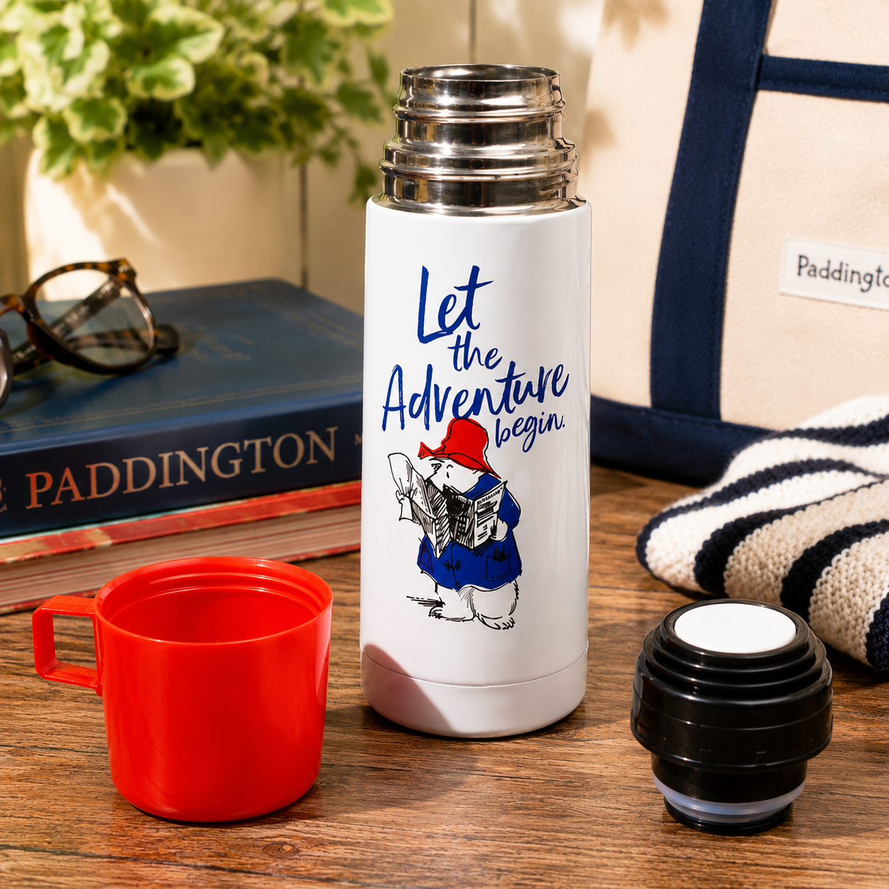 White thermos with Paddington Bear design and text 'Let the Adventure Begin' on a wooden surface with a red cup and black cap.
