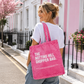 Woman holding a pink tote bag with 'The Notting Hill Shopper Bag' text on a street with pink buildings.