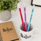 Three Beatles-themed pencils in a mug with a notebook next to it on a white surface.