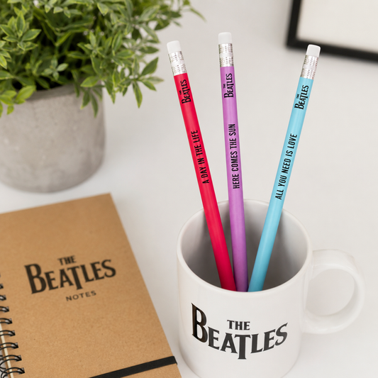 Three Beatles-themed pencils in a mug with a notebook next to it on a white surface.