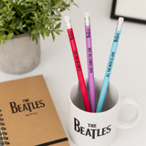 Three Beatles-themed pencils in a mug with a notebook next to it on a white surface.