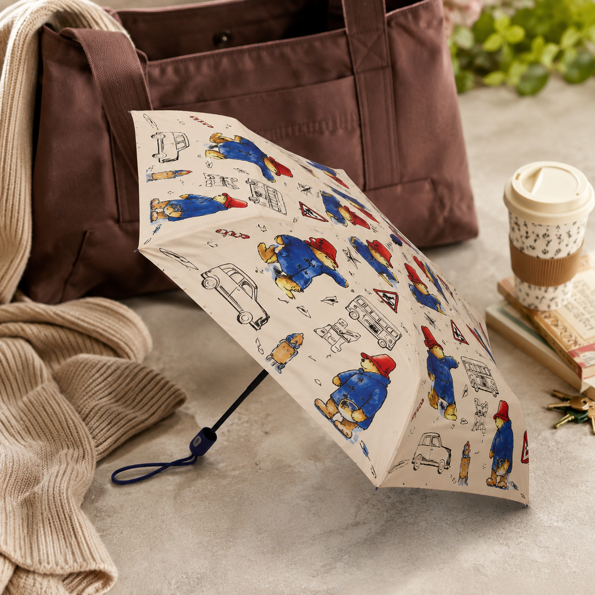 Open Paddington Bear print umbrella displayed indoors beside a brown bag, knitted clothing, books and a takeaway cup.