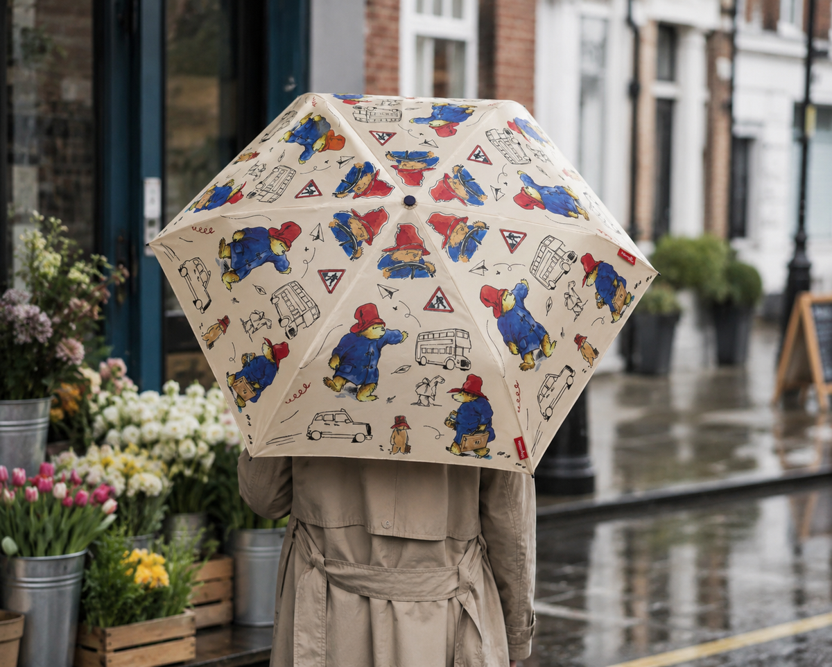 Outside the flower shop:
Person holding an open cream Paddington Bear print umbrella on a wet street beside a flower shop.