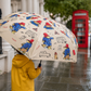 Person in a yellow raincoat holding a cream Paddington Bear print umbrella on a rainy London street, with a red telephone box in the background.
Outside the flower shop: