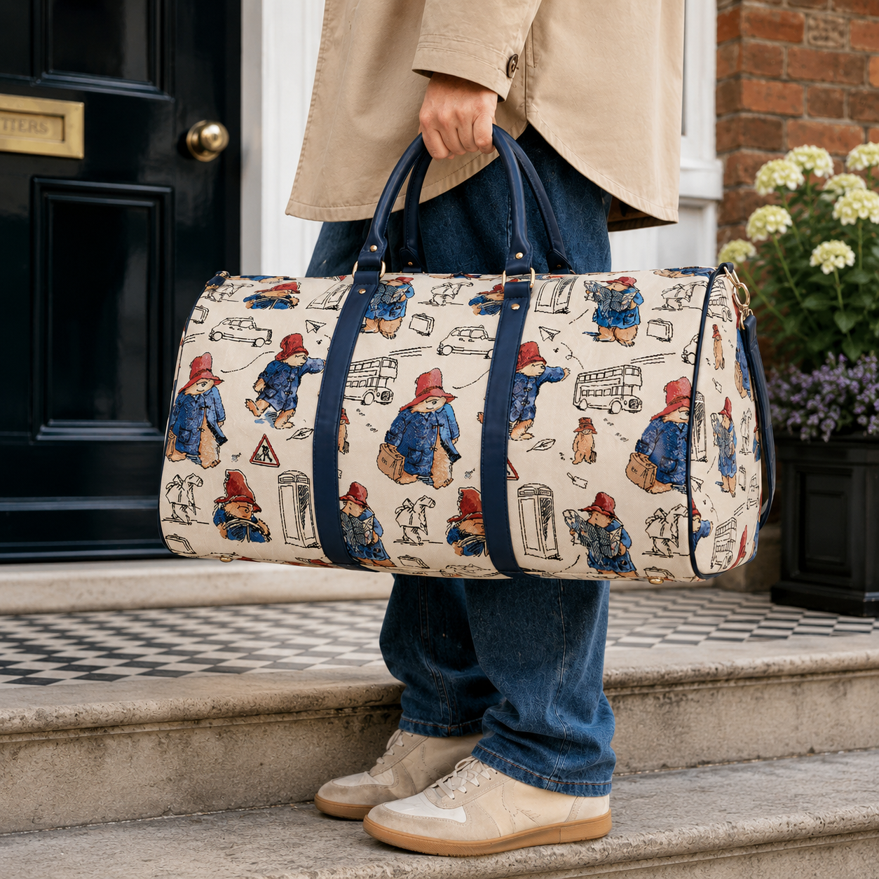 Person carrying a cream Paddington Bear print weekend travel bag with navy handles outside a London townhouse.